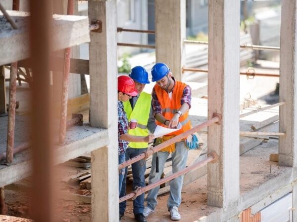 A structural engineer examining a building's framework on-site, holding blueprints with construction workers and equipment in the background